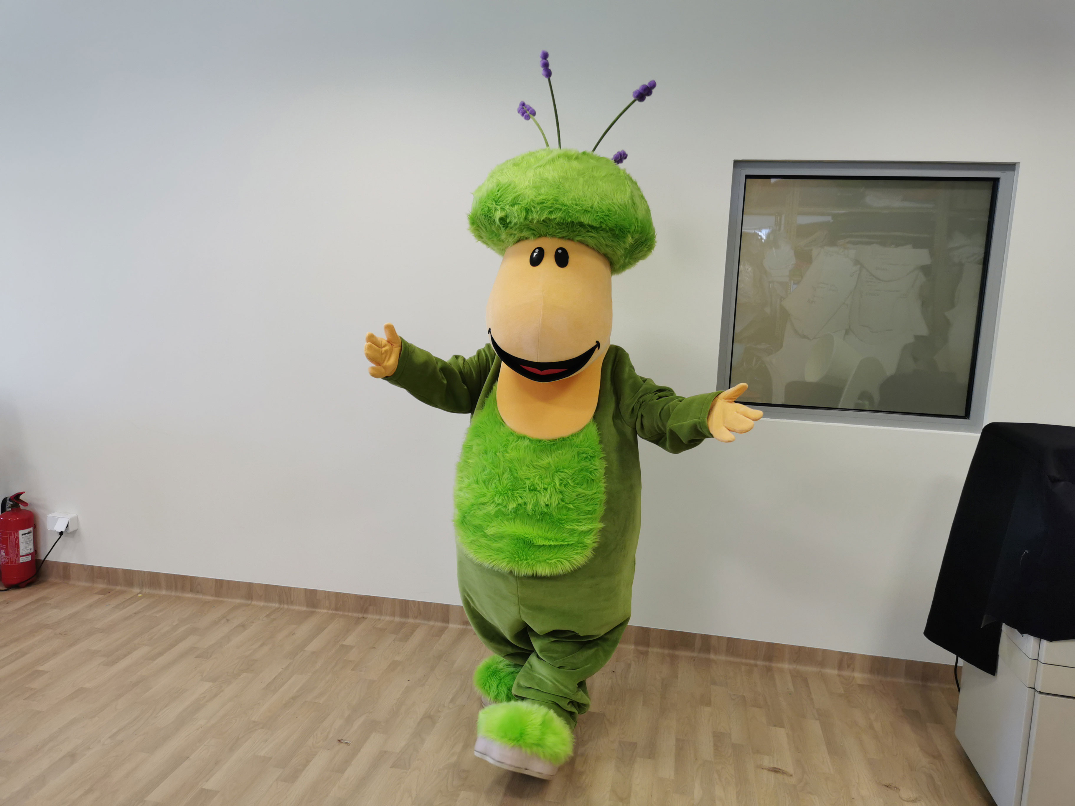 Na fotografii widnieje maskotka ustylizowana na postać zielonego stworka pozująca do zdjęcia w siedzibie firmy Elabika